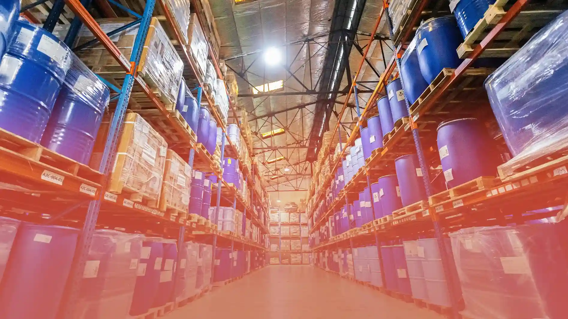 A well-organized Toplis Logistics' warehouse with tall racks storing blue industrial barrels and packaged goods. Bright lighting reflects off the metal ceiling, highlighting the structured storage system.