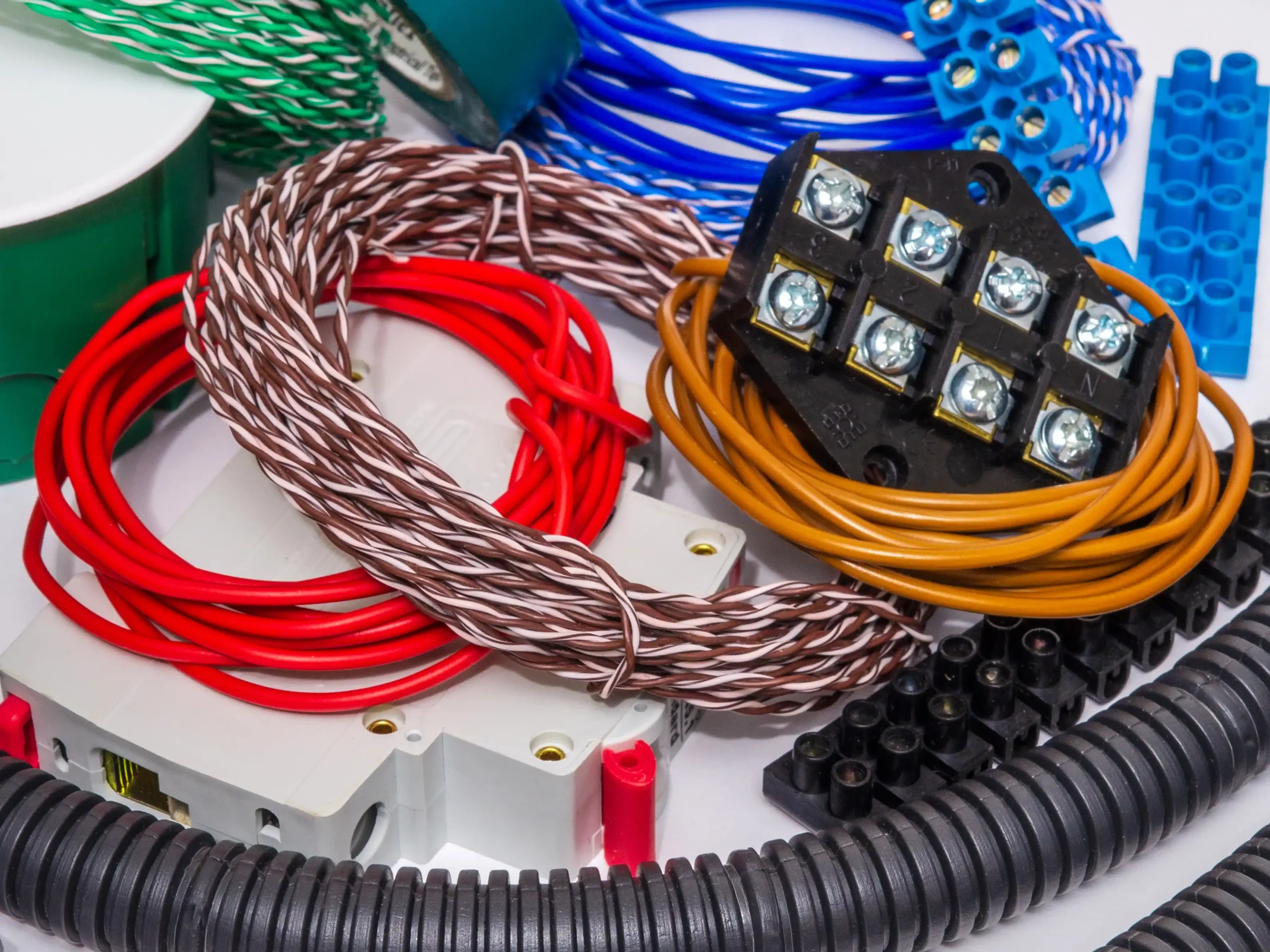 Collection of electrical components including coils of insulated wire in red, brown-and-white twisted pair, orange, blue, and green, black terminal block with screw connectors, gray junction box with red clips, corrugated black conduit tubing, and blue modular connectors