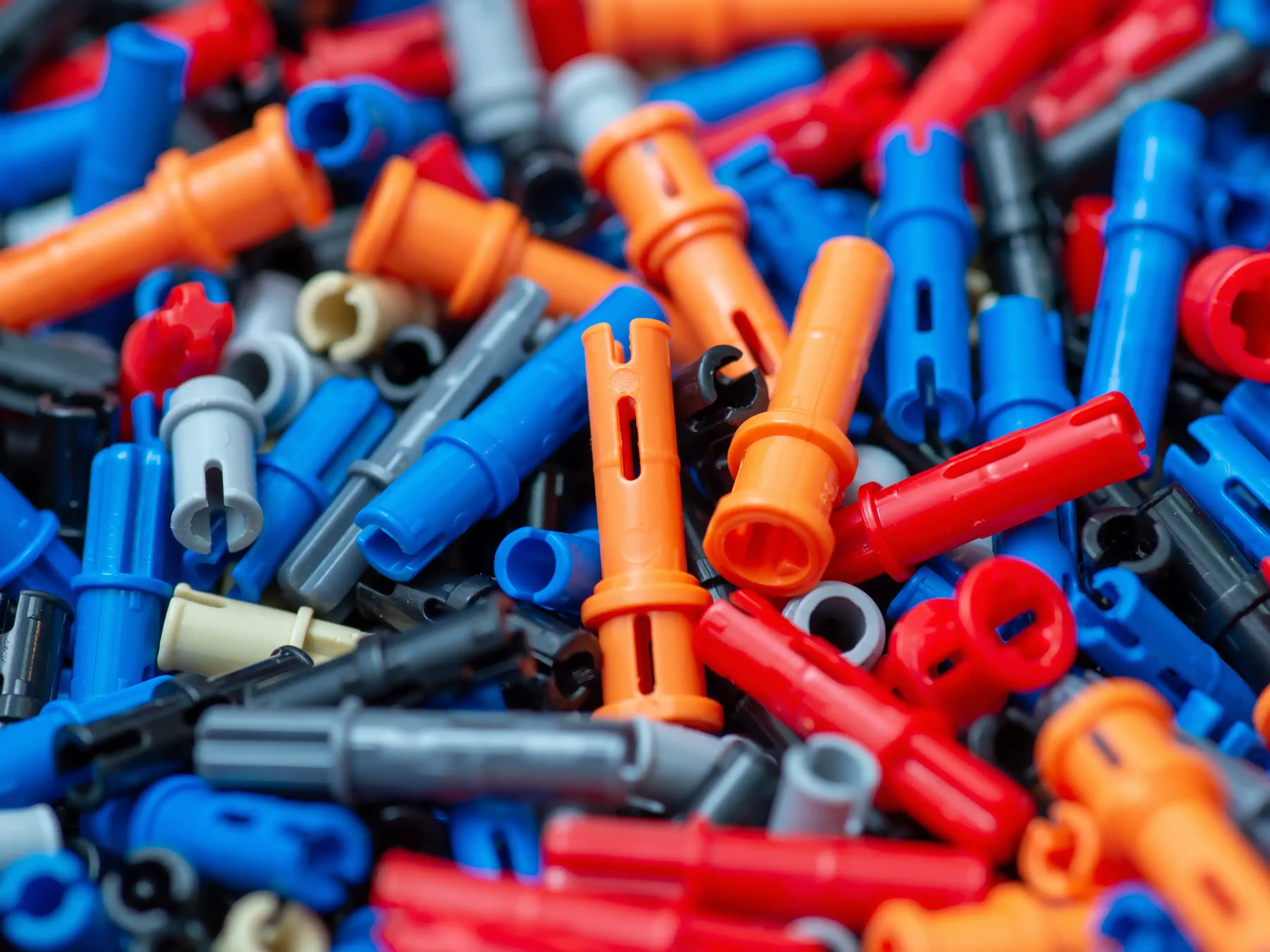 Close-up pile of colorful LEGO Technic connector pins in red, blue, orange, gray, black, and beige with cylindrical shapes and interlocking holes for mechanical builds