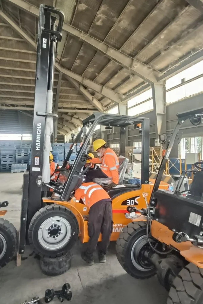 Group of workers in orange safety jackets and yellow helmets performing maintenance on orange HANGCHA forklift model 30 S inside industrial warehouse with high metal-beamed roof and stacks of blue pallets in background