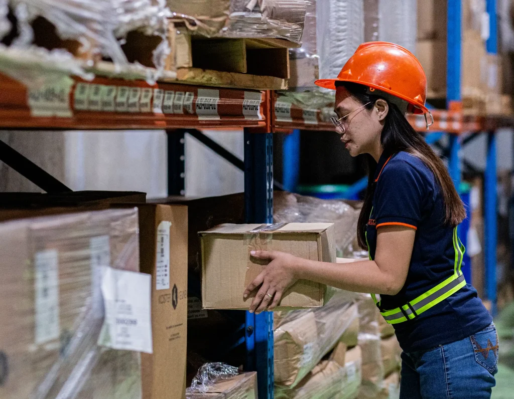 Warehouse worker wearing bright orange safety helmet safety glasses and navy-blue reflective shirt handling cardboard box on organized metal shelving unit filled with labeled packages representing logistics safety and manual handling