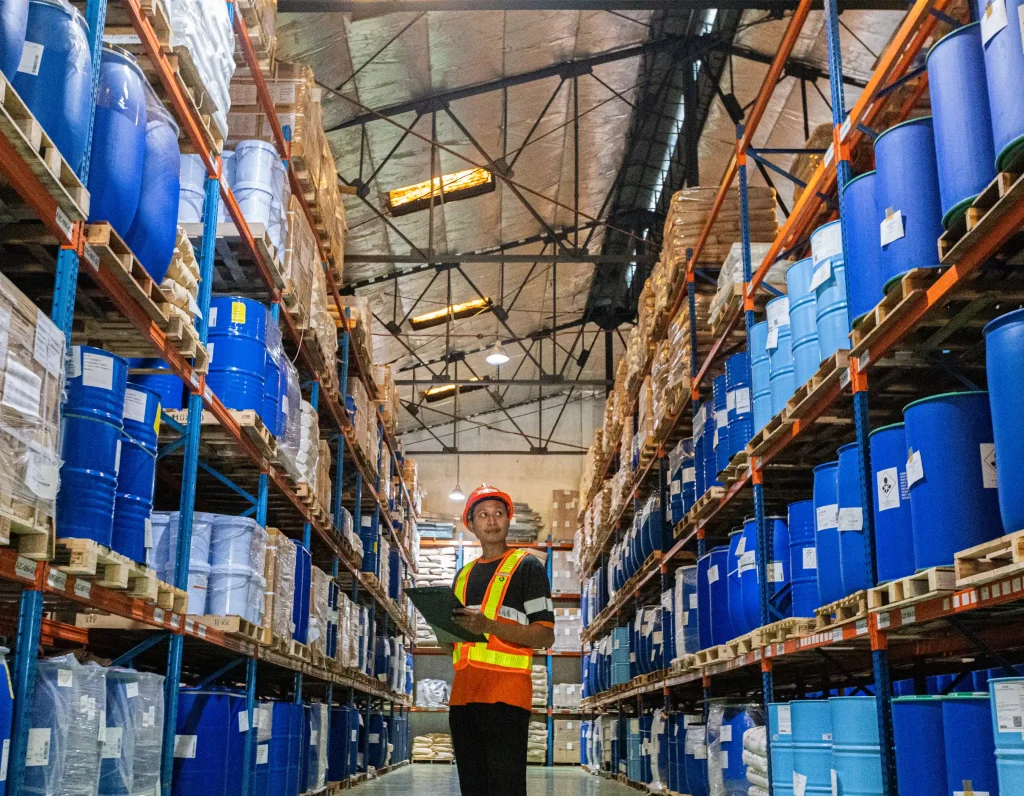 Large warehouse interior with tall metal shelving units stacked with blue barrels and containers, person in safety vest and hard hat holding clipboard inspecting inventory under high ceiling with metal beams and industrial lighting