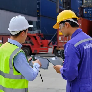 Two workers in safety gear hard hats and reflective vests standing in industrial shipping yard one holding tablet and other with clipboard and pen with stacked shipping containers and red container-handling vehicle in background representing logistics inspection and operations management